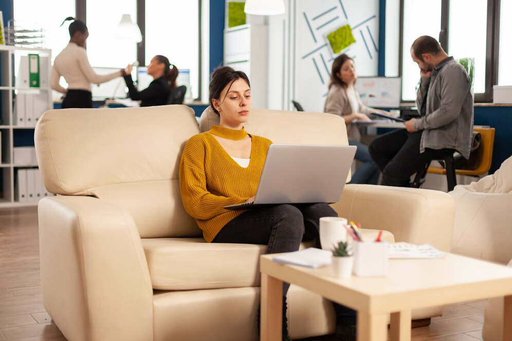 Careers 13 Woman working on a laptop in a modern office lounge with a collaborative team in the background.